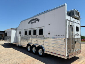 A large white and silver Bloomer gooseneck horse trailer with a 22' loft, three axles, and multiple windows is parked on gravel under a clear blue sky. This 4 Horse Side Load trailer features &ldquo;Bloomer Trailers&rdquo; branding and an air conditioning unit on top. -Stillwater Trailer Sales