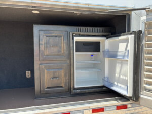 A small refrigerator with its door open, showing empty shelves and door bins, is built into dark wooden cabinets in a horse trailer or 22' loft RV setup. -Stillwater Trailer Sales