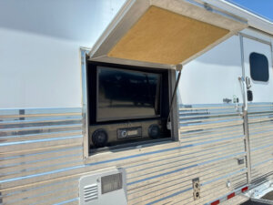 A silver Bloomer horse trailer with a built-in outdoor entertainment system, featuring an open compartment with a TV, two speakers, and a stereo under a lifted hatch. -Stillwater Trailer Sales