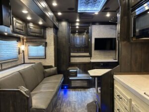 Interior of a modern RV with dark wood cabinets, a gray couch, and a wall-mounted TV. Soft lighting and windows with shades create a cozy, comfortable atmosphere&mdash;perfect for relaxing after hauling with your Bloomer 4 Horse Trailer. -Stillwater Trailer Sales
