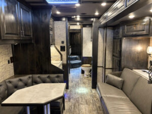 Interior of a modern Bloomer RV with dark wood cabinets, gray upholstered seating, a small dining area, and a kitchen in the background. A visible bathroom with toilet is at the back. The space is well-lit with LED lights and features a 22' loft. -Stillwater Trailer Sales