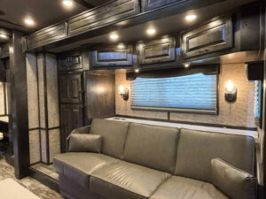 Modern RV living area featuring dark wood cabinets, overhead lighting, a large gray sofa, two pillows, textured wallpaper, wall sconces, and a window with a pleated shade&mdash;perfect for relaxing in your Bloomer 4 Horse side load trailer. -Stillwater Trailer Sales