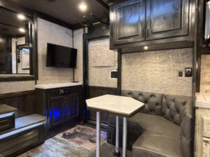 Modern Bloomer 4 Horse RV interior with dark wood cabinets, a small mounted TV, a corner leather bench with marble-top table, and textured walls. Steps with blue accent lighting lead to the side load area. -Stillwater Trailer Sales
