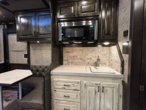 A compact RV kitchen in a Bloomer 22' Loft features dark wood cabinets, a built-in microwave, a white distressed cabinet below, small sink, and light tile backsplash. To the left, a dining area with booth seating overlooks the 4 Horse Side Load area. -Stillwater Trailer Sales