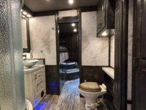 Modern RV bathroom with a glass shower, vessel sink on a white vanity, and brown toilet. Dark wood cabinets contrast light walls. A door opens to a cozy bunk area&mdash;perfect for a Bloomer horse trailer with convenient side load access and seating. -Stillwater Trailer Sales