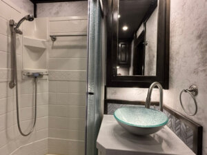 Modern bathroom with a glass vessel sink on a white countertop, a round mirror above, and a walk-in shower&mdash;an upscale touch in this Bloomer 4 Horse Side Load with 22' Loft living quarters. -Stillwater Trailer Sales