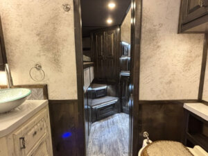 A view from a small bathroom with a patterned sink, vanity, and toilet, looking through a doorway into the wood-paneled hallway of this Bloomer 22' Loft with steps leading up to cabinets and a cozy loft area. -Stillwater Trailer Sales