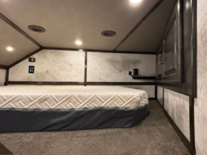 A small, carpeted room with a low ceiling, reminiscent of a cozy 22' Loft, featuring a mattress on the floor, a shelf with a few items, wall outlets, and recessed lighting. The textured walls enhance the compact and inviting atmosphere. -Stillwater Trailer Sales