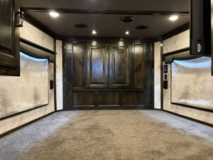 A wide, carpeted room with dark wooden cabinets at the far end, white textured walls, recessed ceiling lights, and built-in shelving on both sides&mdash;reminiscent of the luxurious interiors found in a Bloomer horse trailer. -Stillwater Trailer Sales