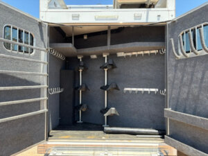 The image shows the open rear storage compartment of a Bloomer 4 horse trailer, featuring multiple hooks, saddle racks, and carpeted walls for organizing tack and equipment, all illuminated by sunlight. -Stillwater Trailer Sales