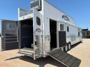 A large Bloomer 4 Horse Side Load trailer with open doors and ramps, revealing interior stalls, storage hooks, and a spacious 22' Loft, is parked on a gravel lot under a clear blue sky. -Stillwater Trailer Sales