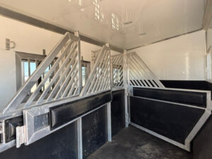The image shows the interior of a Bloomer 4 horse trailer with empty, divided stalls. The stalls are separated by metal bars and padded dividers, with a side load design and white walls featuring black padding along the lower half. -Stillwater Trailer Sales