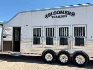 A large white Bloomer 4 Horse Side Load trailer with three axles, several barred windows, and an open door is parked on gravel under a clear blue sky. -Stillwater Trailer Sales