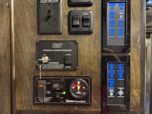 Control panel with switches, buttons, digital displays, and indicator lights for functions like room extension, generator control, and lighting in a Bloomer 22' Loft 4 Horse Side Load trailer, mounted on a wood-paneled wall. -Stillwater Trailer Sales