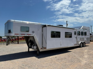 A large white Featherlite 4 Horse Trailer with living quarters is parked on gravel under a partly cloudy sky. The trailer features multiple windows, a slide-out section, and two axles. -Stillwater Trailer Sales