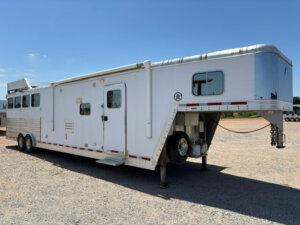 A large white Featherlite 4 Horse gooseneck trailer with windows, side door, retractable awning, and spacious living quarters is parked on gravel under a clear blue sky. -Stillwater Trailer Sales