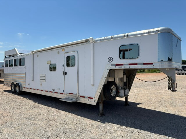 A large white Featherlite 4 Horse Trailer with living quarters is parked on gravel under a clear blue sky. The trailer features multiple windows, ample storage compartments, and two sturdy axles. -Stillwater Trailer Sales