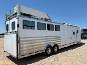 A large white Featherlite gooseneck trailer with several windows, dual axles, rooftop storage, and spacious living quarters sits parked on gravel under a clear blue sky. -Stillwater Trailer Sales