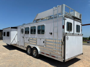 A large silver Featherlite 4 Horse trailer with living quarters features multiple windows, double axles, an external ladder, roof rack, and a side slide-out, parked on gravel under a clear blue sky. -Stillwater Trailer Sales