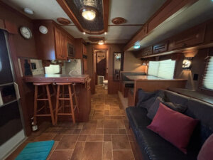 Step inside the Featherlite 4 Horse Trailer&rsquo;s living quarters, featuring wood cabinetry, a compact kitchen with bar and stools, a black sofa with pillows, booth-style dining area, and a hallway leading to the rear bathroom filled with natural light. -Stillwater Trailer Sales