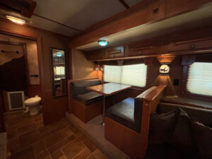 A cozy RV interior in this Featherlite 4 Horse Trailer features a dining booth with dark cushions, wood accents, overhead cabinets, and two windows with blinds. To the left, a door and toilet are visible. The living quarters are warm and inviting. -Stillwater Trailer Sales