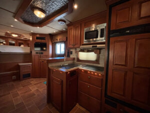 Interior of a Featherlite 4 Horse RV kitchen featuring wood cabinets, a sink, stove, microwave, refrigerator, and built-in TV. Under-cabinet lighting and a small window above the sink make these living quarters compact and warmly lit. -Stillwater Trailer Sales