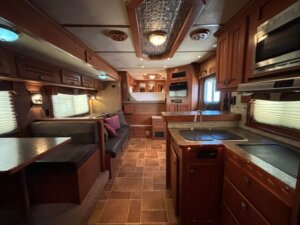 Interior of cozy Featherlite 4 Horse living quarters, featuring wood cabinets, a small kitchen with sink and microwave, a dining area with bench seating, a sofa, and windows with blinds, all warmly lit by overhead lights. -Stillwater Trailer Sales