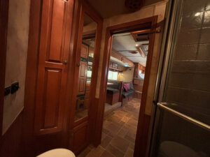 View from a Featherlite 4 Horse Trailer bathroom with wooden cabinets and a glass shower door, looking into cozy living quarters with tiled floors, seating, and windows with blinds. Warm lighting creates an inviting atmosphere. -Stillwater Trailer Sales