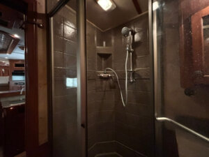 A glass-enclosed, corner shower with dark brown tile walls, a mounted showerhead with hose, and a built-in soap dish inside this Featherlite 4 Horse Trailer&rsquo;s RV bathroom. Kitchen and living quarters are visible through an open doorway. -Stillwater Trailer Sales