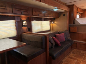 A cozy Featherlite RV interior with wooden cabinets, a cushioned bench dining area, a small sofa with pillows, and a bed area in the background. Warm lighting and closed blinds create a comfortable atmosphere in the living quarters. -Stillwater Trailer Sales