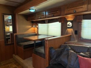 A cozy Featherlite RV interior features a wooden dining booth with black cushions, a mirrored wall, overhead storage cabinets, a cushioned sofa with pillows, and blinds covering the windows. Warm lighting enhances these inviting living quarters. -Stillwater Trailer Sales