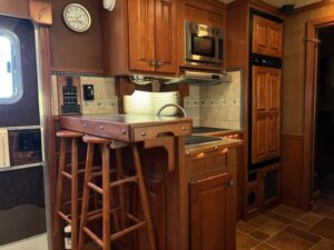 A cozy Featherlite 4 Horse Trailer kitchen features wooden cabinets, a built-in microwave, stovetop, refrigerator, and a small counter with two bar stools. Soft string lights line the counter, creating inviting living quarters. -Stillwater Trailer Sales