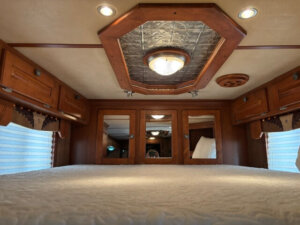 Interior view of a Featherlite 4 Horse Trailer bedroom with a large bed, wood-paneled cabinets, mirrors on the back wall, and a decorative ceiling light fixture. Soft lighting in the living quarters creates a cozy atmosphere. -Stillwater Trailer Sales