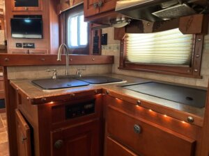 A compact Featherlite RV kitchen with wood cabinetry, a double sink covered by lids, a covered stovetop, and under-cabinet lighting. Perfect for living quarters, it features a window with blinds above the sink and a mounted TV in the background. -Stillwater Trailer Sales