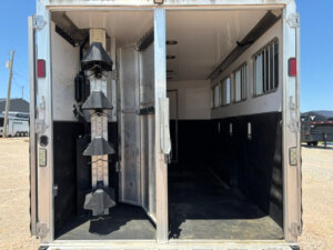 The image shows the open rear view of a Featherlite horse trailer with two separate sections: a tack room on the left with saddle racks and a larger 4 horse compartment on the right with rubber flooring and windows. -Stillwater Trailer Sales