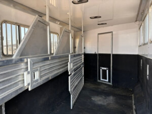 Interior of an empty Featherlite 4 Horse Trailer with metal dividers, barred windows, and a small rear door. The black and white walls and the dark non-slip floor add both style and safety to its spacious living quarters. -Stillwater Trailer Sales