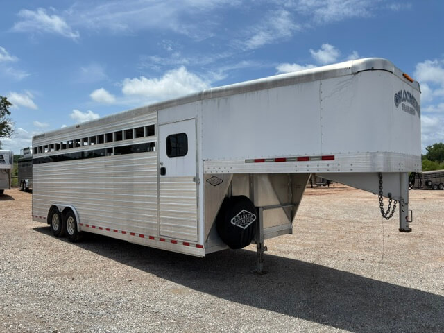 A large silver Bloomer Stock Combo livestock trailer, measuring 24', with a gooseneck hitch is parked on a gravel lot under a partly cloudy sky. The trailer features multiple vents along the side and is supported by two axles with dual wheels. -Stillwater Trailer Sales