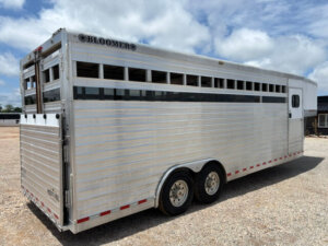 A large 24' silver Bloomer Stock Combo livestock trailer with dual axles is parked on a gravel lot under a partly cloudy sky. The trailer features horizontal vents and a rear gate. -Stillwater Trailer Sales