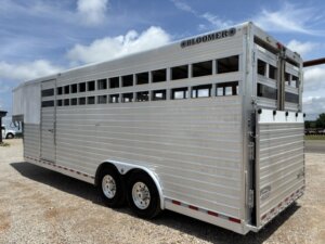 A large silver Bloomer Stock Combo livestock trailer, 24' in length, with ventilation slats and dual axles, is parked on gravel under a partly cloudy sky. -Stillwater Trailer Sales