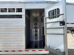 Open door on a silver 24 Foot Trailer reveals a clean, empty tack room with saddle racks and bridle hooks inside. This Stock Combo Trailer features aluminum siding and barred windows, parked on a gravel lot. -Stillwater Trailer Sales
