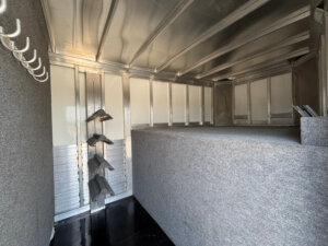 Interior of a Bloomer Trailer with metal walls and ceiling, grey carpeted partitions, saddle racks on the left wall, and hooks for hanging equipment. The space is clean and well-organized. -Stillwater Trailer Sales