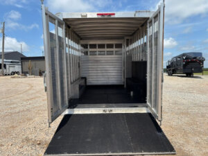 Open 24' Bloomer Stock Combo livestock trailer with ramp down, showing an empty interior with side windows and partitions, parked on gravel under a partly cloudy sky. Other trailers and buildings are visible in the background. -Stillwater Trailer Sales