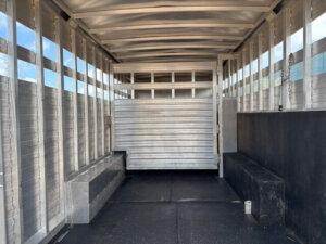Interior of a 24 foot Bloomer trailer stock combo with metal walls, vented sides, a central dividing gate, and non-slip black flooring. Benches line both sides beneath the windows while light filters in through vent slats. -Stillwater Trailer Sales
