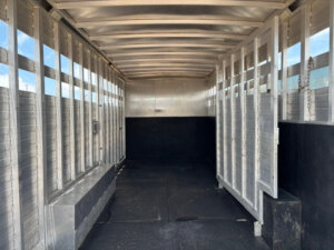 Interior view of an empty Bloomer horse trailer with metal walls, barred sides, and a rubber-matted floor; sunlight streams through the slats, casting shadows inside this stock combo trailer. -Stillwater Trailer Sales