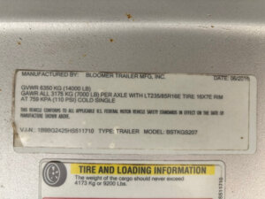 Close-up of a Bloomer Stock Combo vehicle information label showing manufacturer details, weight ratings, tire specifications, VIN, and 24' trailer model. The slightly worn label is affixed to a metallic surface. -Stillwater Trailer Sales