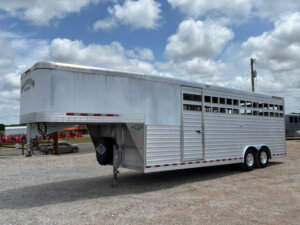 A large silver Bloomer 24 Livestock Trailer with slatted sides is parked on gravel under a partly cloudy sky. This Stock Combo Trailer has two axles and is designed for transporting animals. -Stillwater Trailer Sales