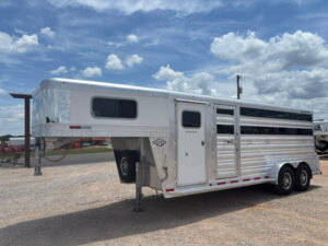 A Platinum Stock Combo 20' silver gooseneck livestock trailer with two axles is parked on a gravel lot under a partly cloudy sky. The trailer features side vents, a white door, and its front end elevated on a support jack. -Stillwater Trailer Sales