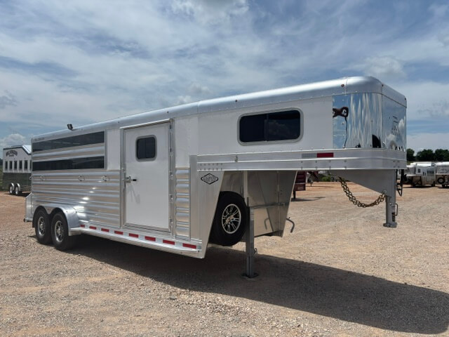 A shiny Platinum 20' gooseneck horse trailer with windows and a side door is parked on a gravel lot under a partly cloudy sky. -Stillwater Trailer Sales