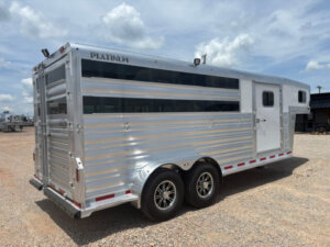 A shiny, silver Platinum Stock Combo trailer, 20' long with black-tinted windows and dual axles, is parked on a gravel lot under a partly cloudy sky. The side door and ventilation slots are visible. -Stillwater Trailer Sales