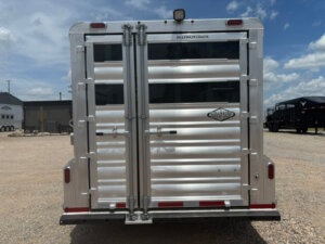 The image shows the rear view of a silver Platinum Stock Combo livestock trailer, 20&rsquo; in length, with two closed doors, metal hinges, and a small Platinum Coach logo. The trailer is parked on gravel under a partly cloudy sky. -Stillwater Trailer Sales